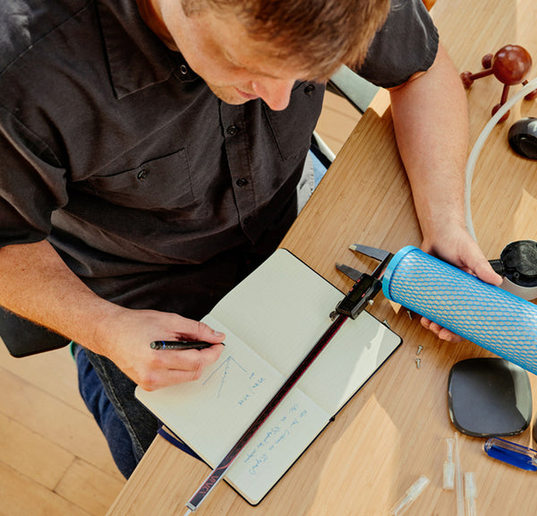 A photo of an individual seated at a desk. He is taking notes with a Hydroviv drinking water refill filter in hand.